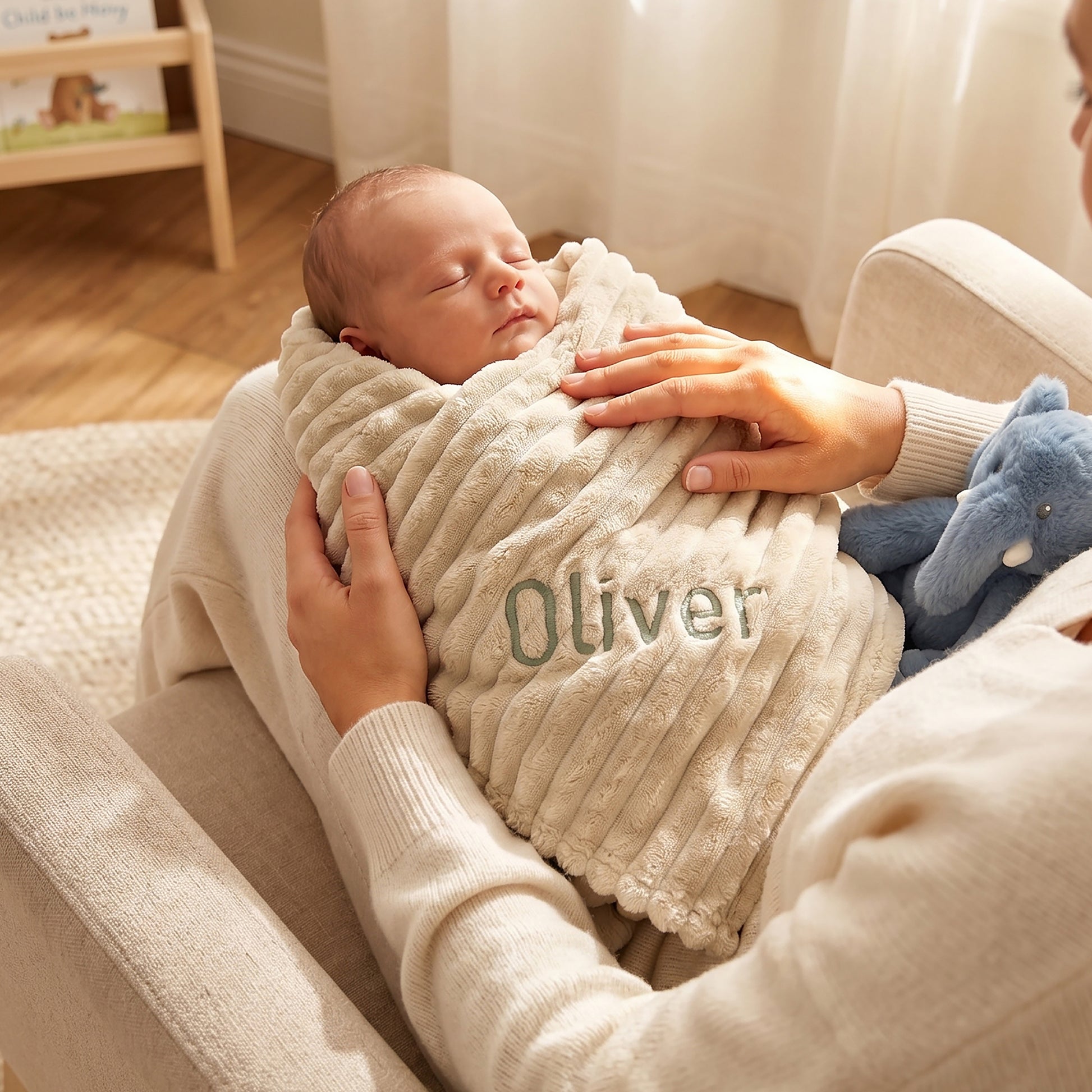 A sleeping newborn baby is swaddled in a cream-colored ribbed blanket with the name 'Oliver' embroidered on the front, held by an adult in a cozy living room setting.