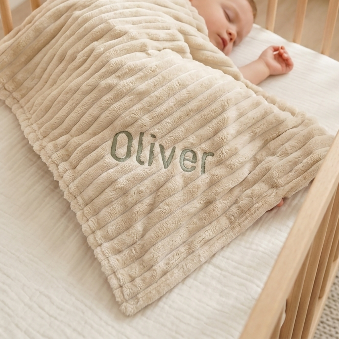 A baby sleeping in a wooden crib, covered with a beige ribbed plush blanket personalized with the name 'Oliver' in green embroidery.