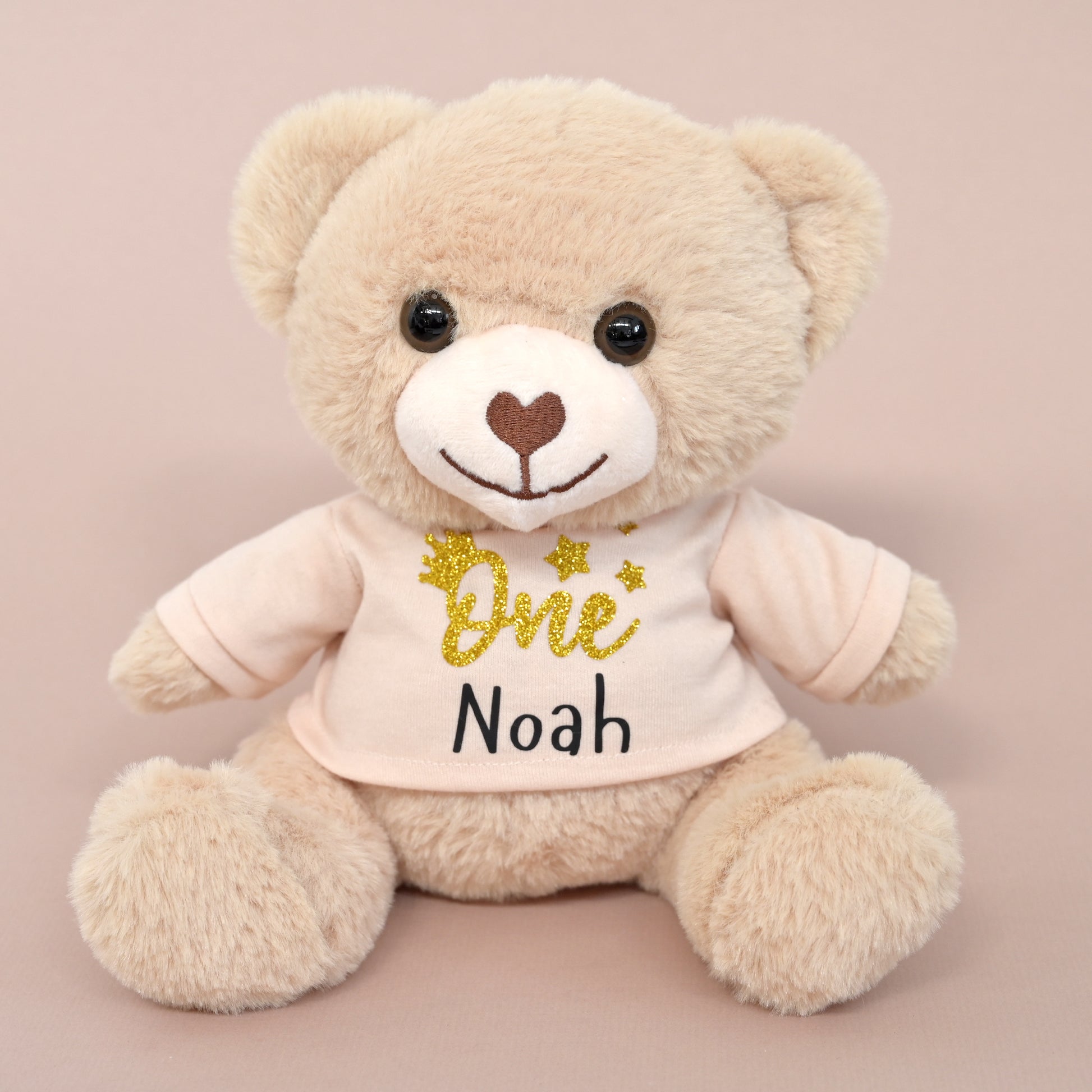 A plush teddy bear in light beige sits against a soft pink background, wearing a pale pink shirt with 'One Noah' printed in black and gold glitter.