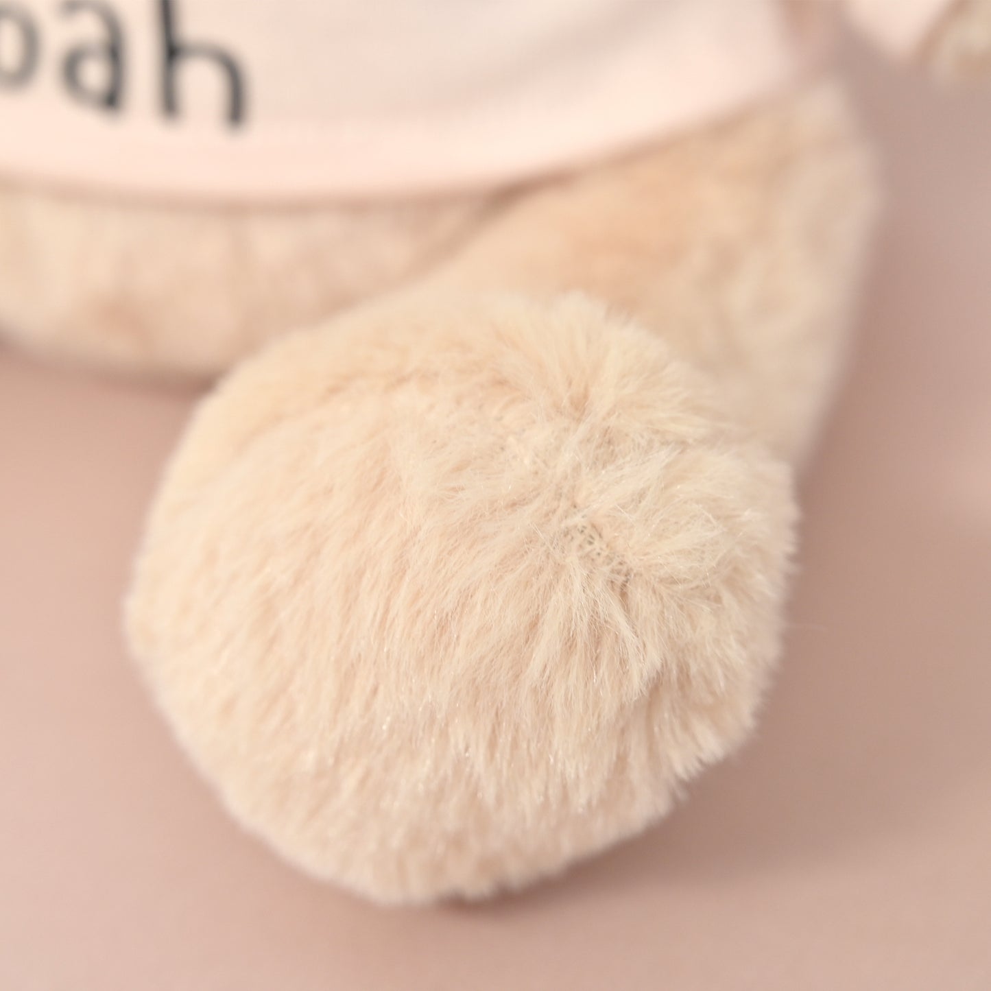 Close-up of a soft plush toy or stuffed animal leg in a warm cream or beige tone, with part of a pale pink body and black lettering visible in the blurred background.