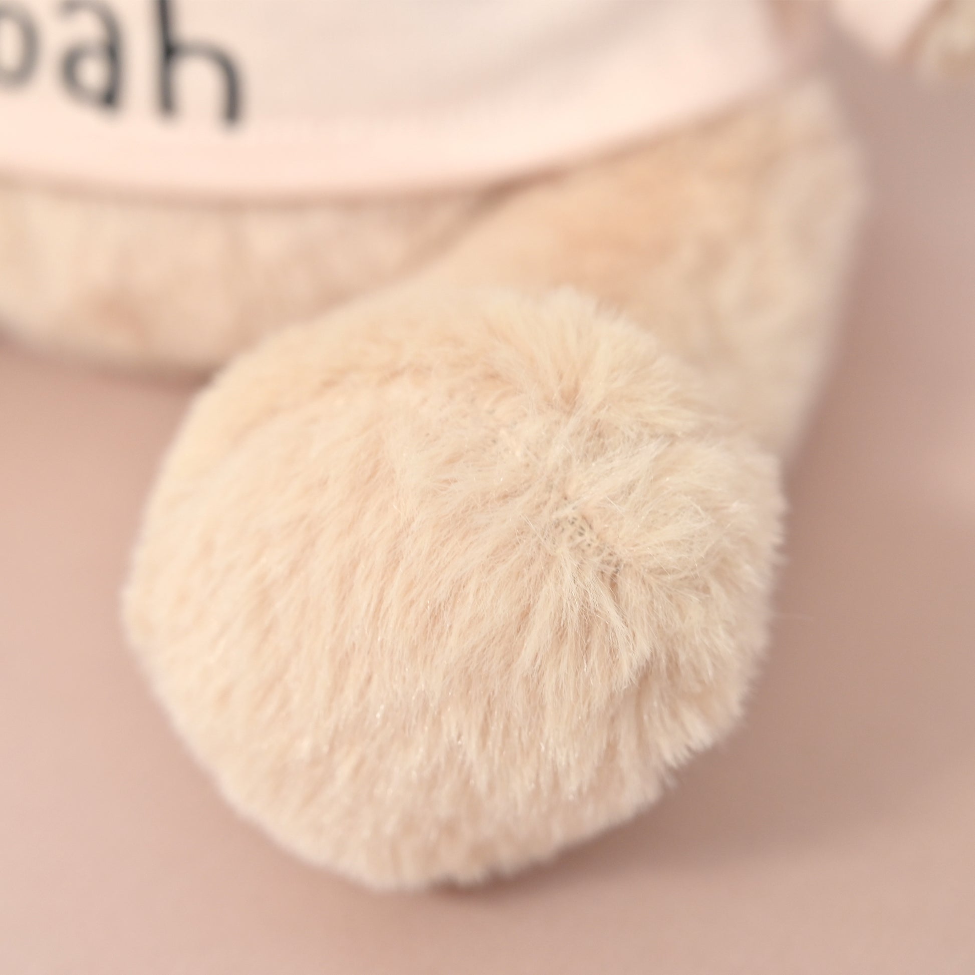 Close-up of a soft plush toy or stuffed animal leg in a warm cream or beige tone, with part of a pale pink body and black lettering visible in the blurred background.
