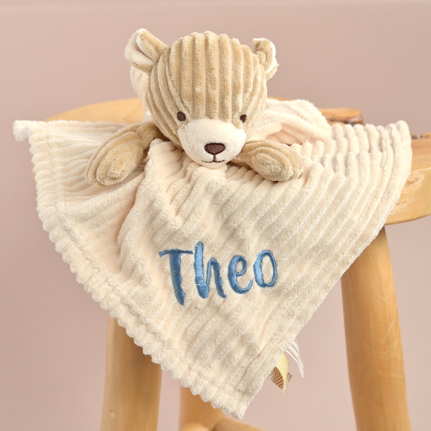 A personalized baby lovey blanket featuring a beige corduroy teddy bear and a cream-colored ribbed fabric cover with the name 'Theo' embroidered in blue, displayed on a wooden stool.