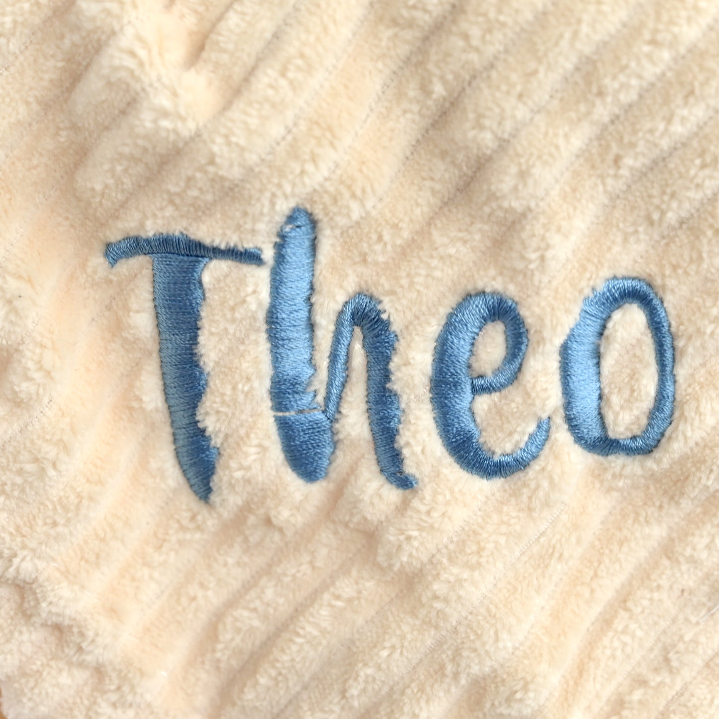 A close-up of a cream-colored plush blanket with textured vertical ribbing, featuring the name 'Theo' embroidered in blue thread.