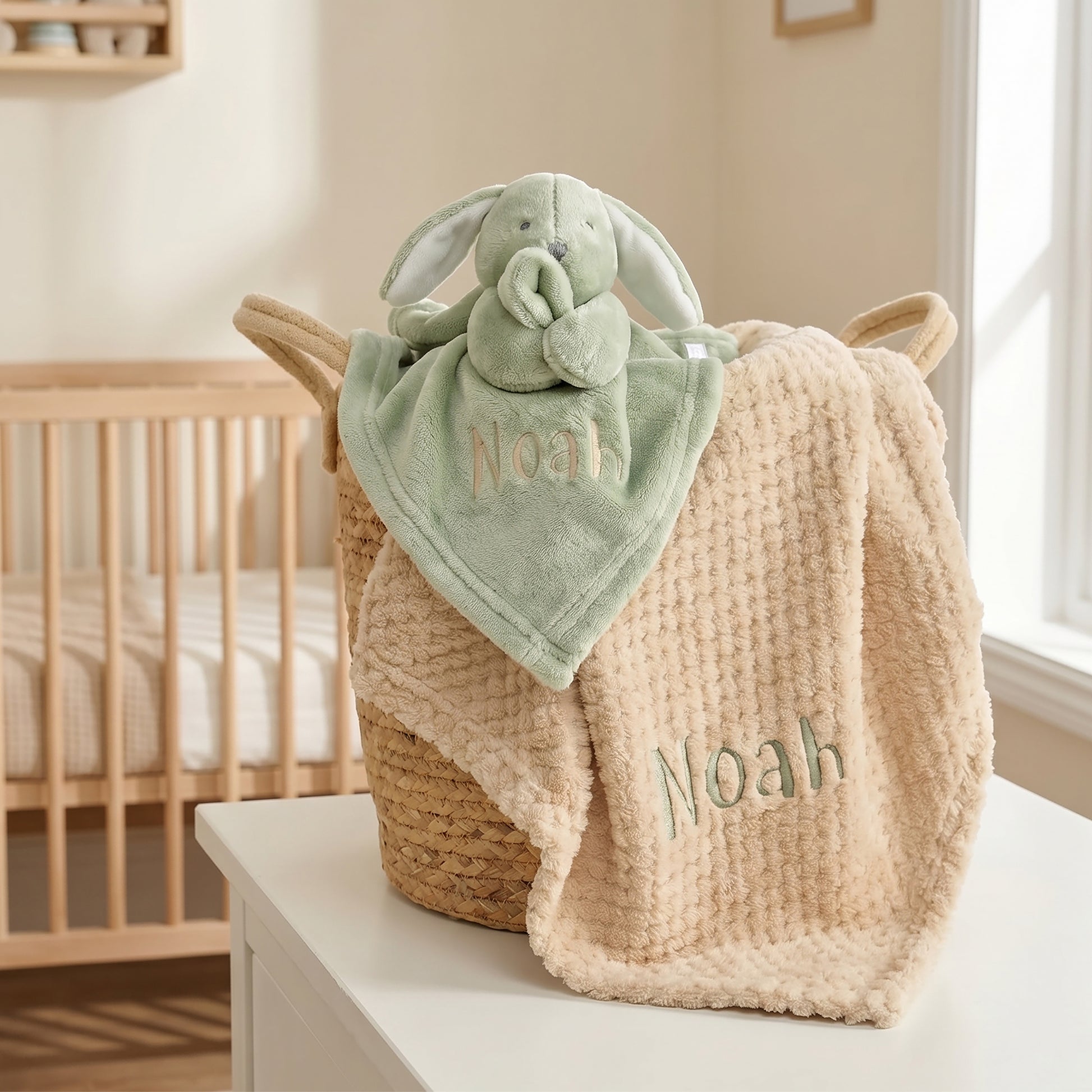 A personalized baby blanket set featuring a textured cream waffle-knit blanket and a soft sage green plush bunny lovey, both embroidered with the name 'Noah' and displayed in a woven basket in a nursery setting.