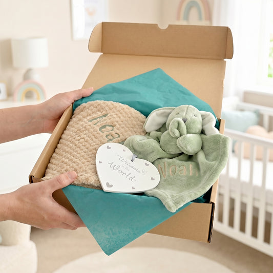 A gift box held by two hands in a softly lit nursery contains a beige knitted blanket with 'Moo' embroidered on it, a plush green elephant toy, a green cloth with 'Mooat' embroidered on it, and a white heart tag that reads 'The Smoother World'.
