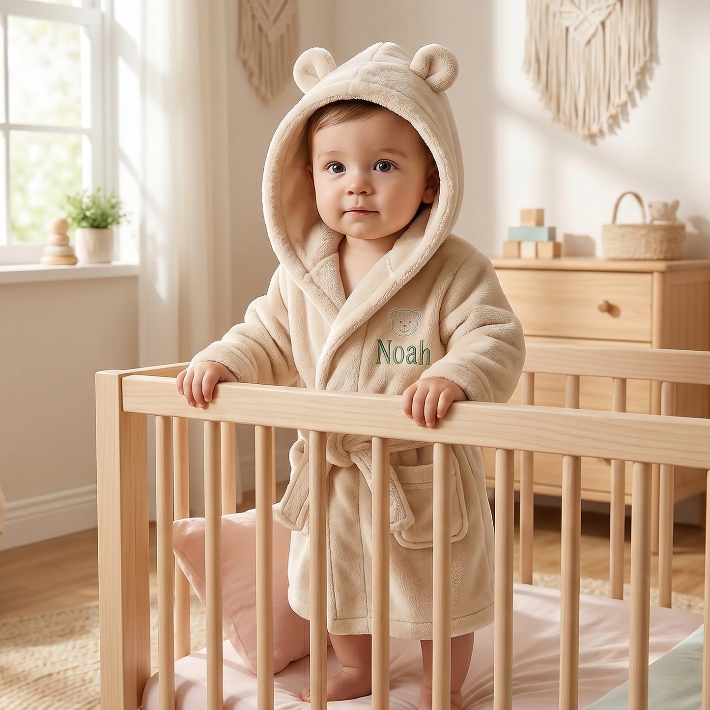 A baby wearing a plush cream-colored hooded robe with bear ears stands in a light wood crib in a bright nursery. The baby's name, 'Noah', is embroidered on the chest.