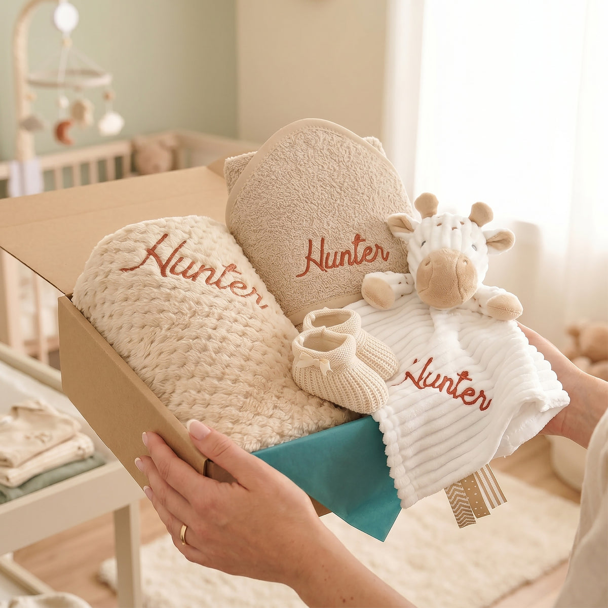 A baby gift set in a cardboard box being held by hands, featuring a chunky knit blanket, a plush giraffe toy, a soft white blanket with 'Hunter' embroidered in brown, and a pair of cream booties, set against a softly blurred nursery background.