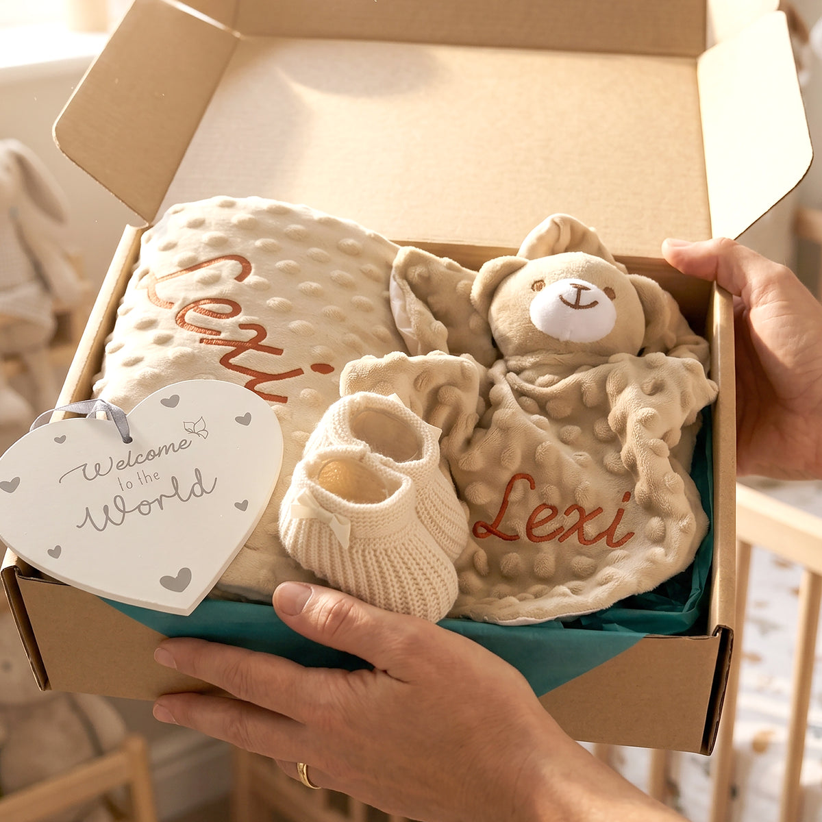 A personalized baby gift box held by hands, containing a cream textured blanket with 'Lexi' embroidered in red, a matching teddy bear toy, a cream knit bootie, and a white heart-shaped 'Welcome to the World' tag.