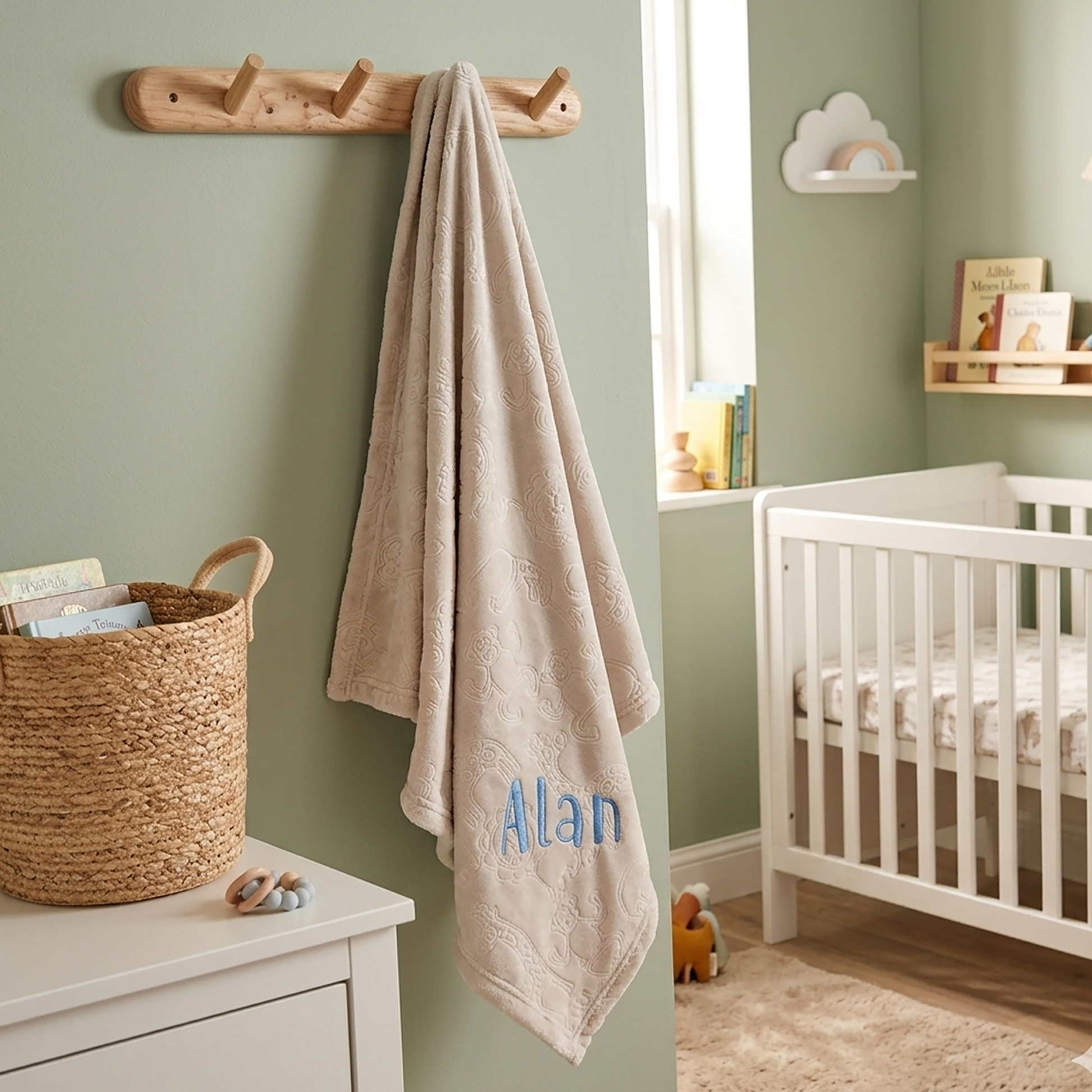 A personalized beige cotton bath towel with the name 'Alan' embroidered in blue hangs from a wooden peg rack on a sage green wall in a styled nursery setting.