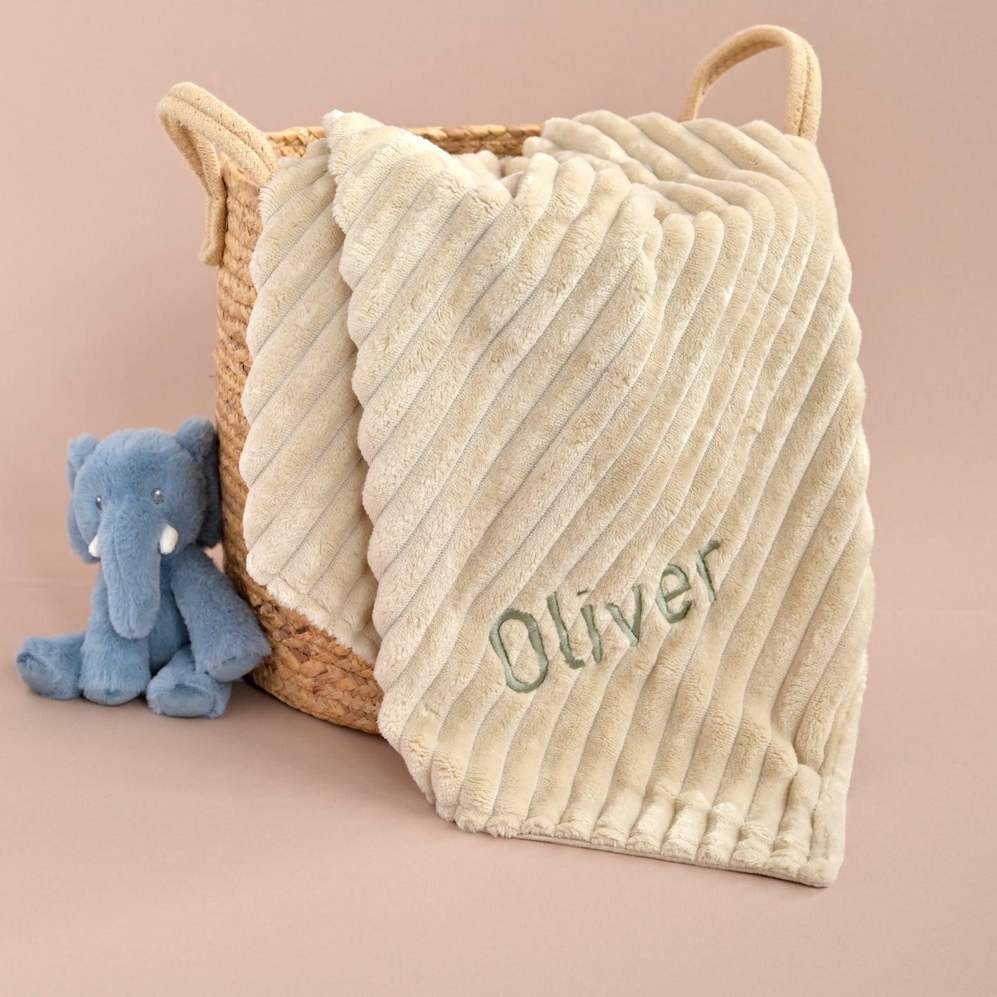 A cream-colored ribbed plush baby blanket with the name 'Oliver' embroidered in green, draped over a woven straw basket next to a small blue plush elephant toy, set against a solid dusty pink background.