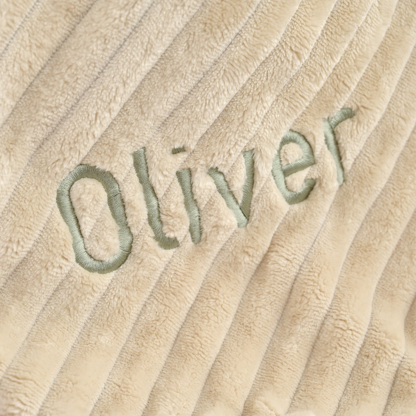 Close-up of a beige ribbed plush fabric with the name 'Oliver' embroidered in light green across the center.
