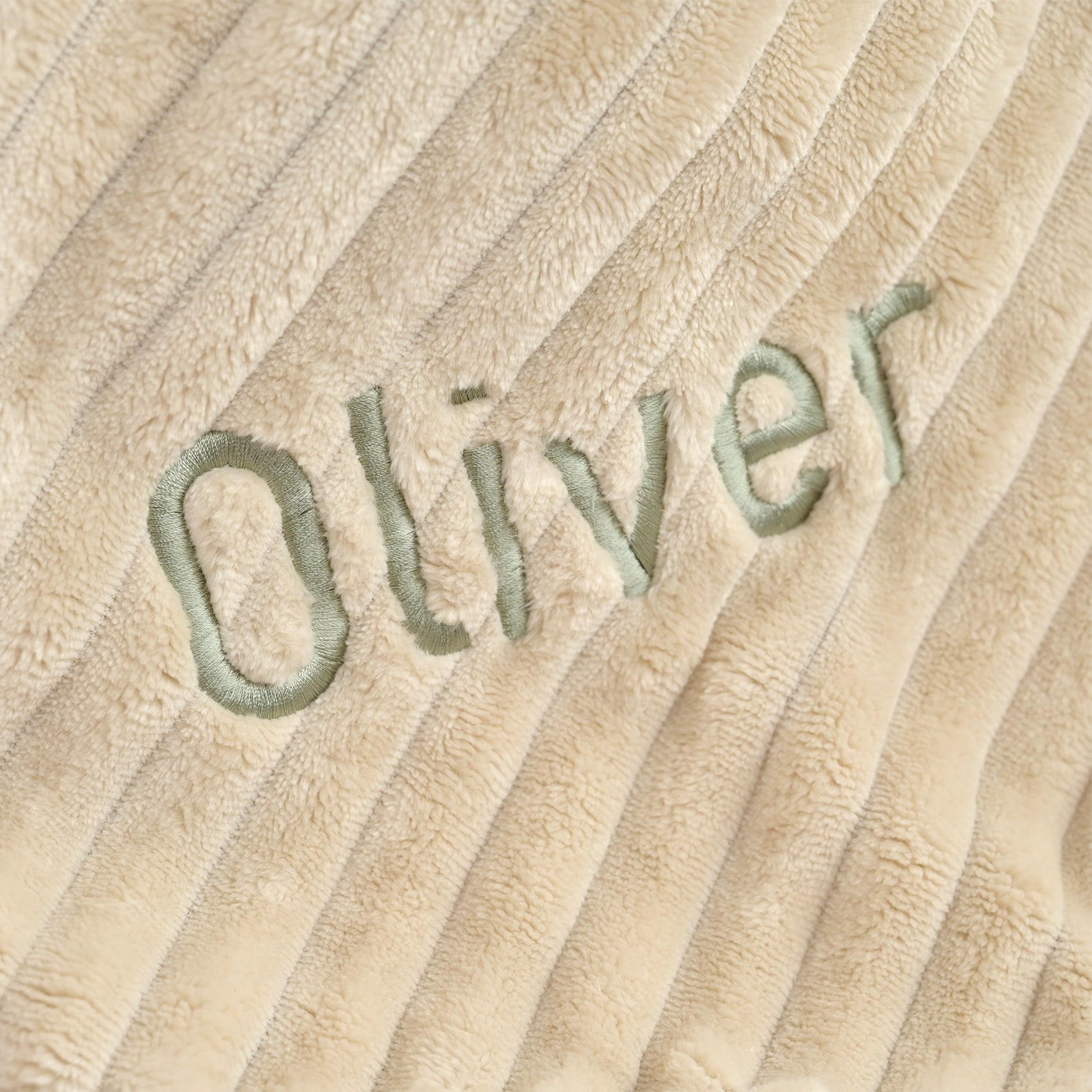 Close-up of a beige ribbed plush fabric with the name 'Oliver' embroidered in light green across the center.