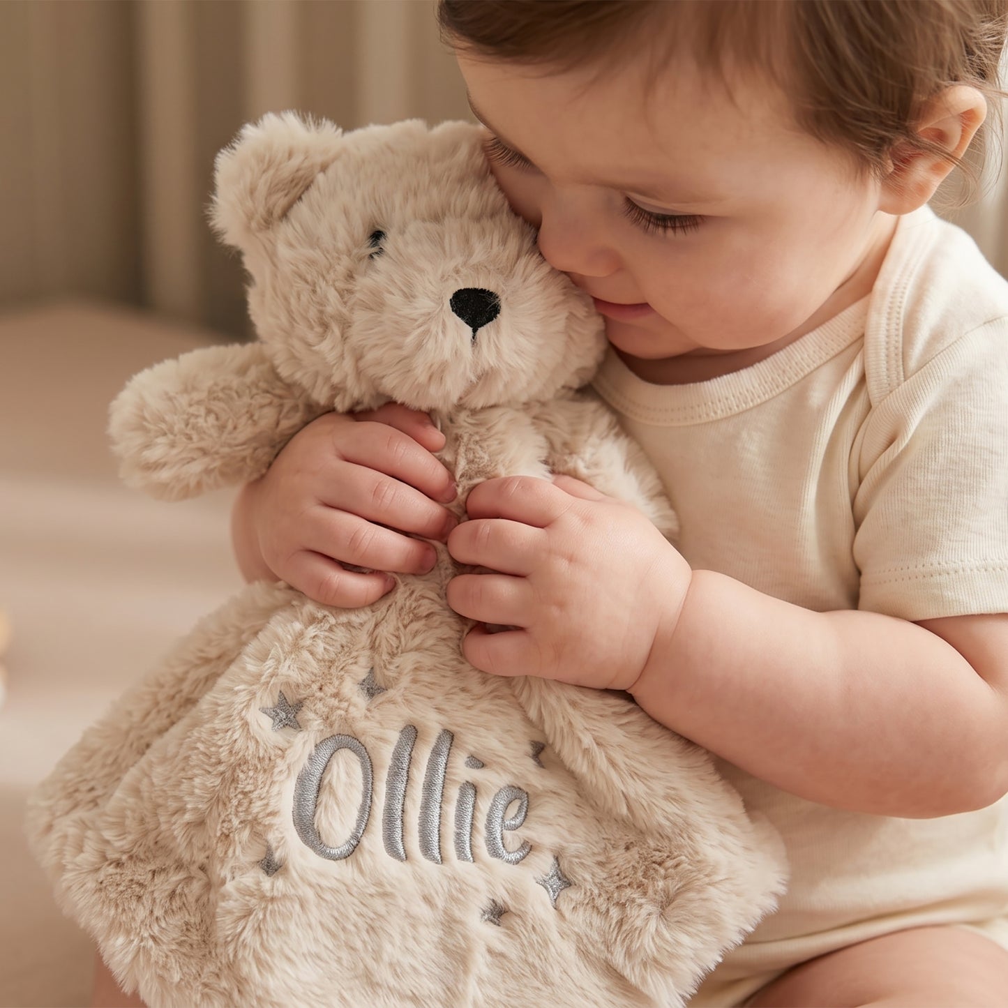 A baby in a cream onesie hugs a plush teddy bear comforter blanket with the name 'Ollie' embroidered on the front, set against a soft, warm indoor background.