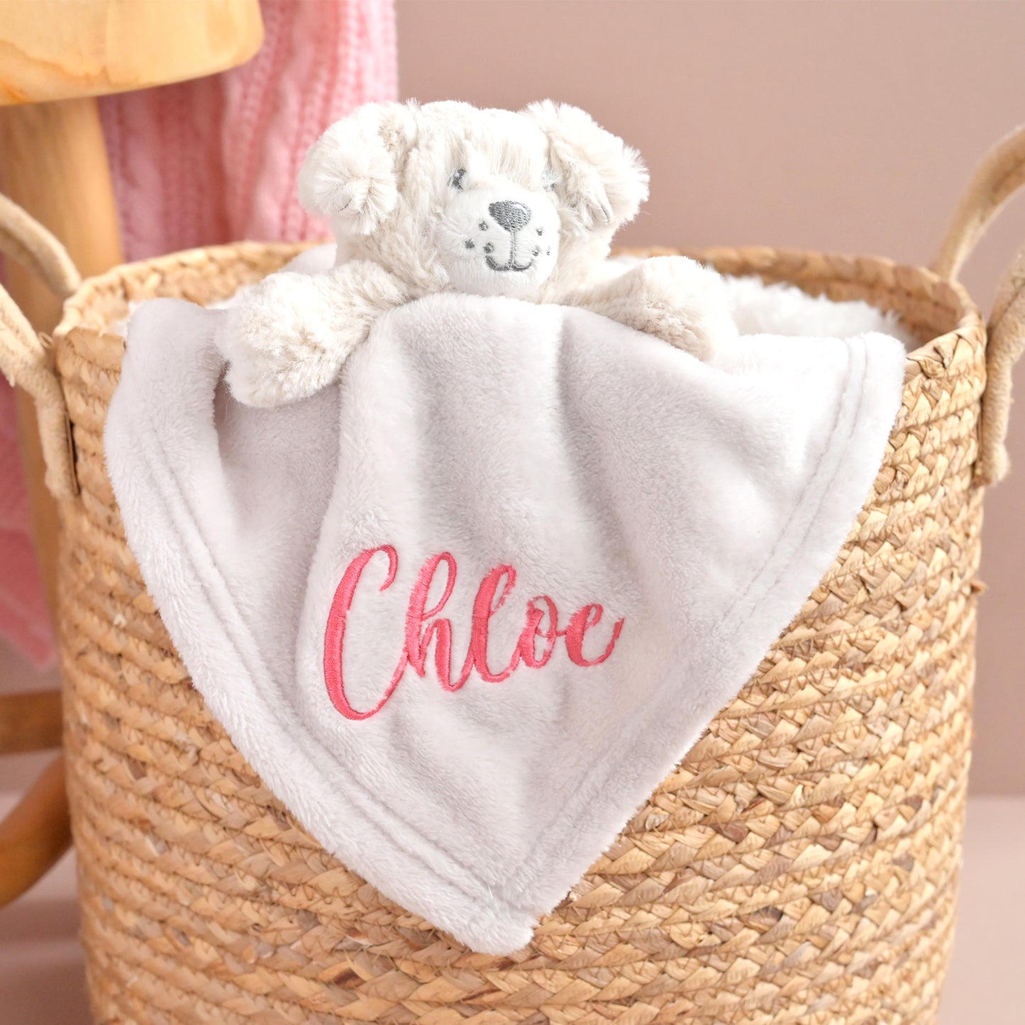 A personalized baby security blanket featuring a light gray plush fabric body with the name 'Chloe' embroidered in pink script, tucked inside a woven straw basket next to a white teddy bear.