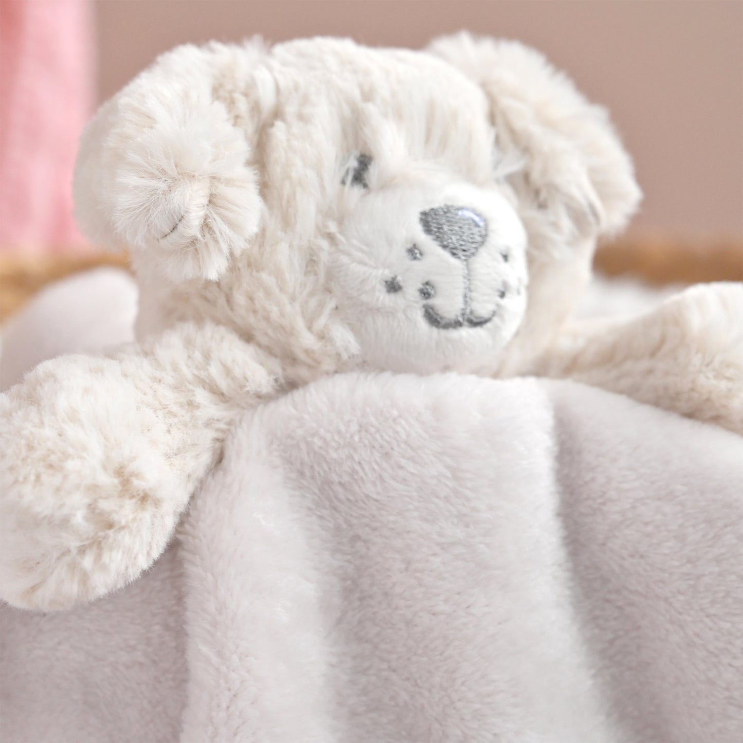 A close-up of a soft plush teddy bear in warm off-white or cream, resting on a matching cream blanket. The bear has embroidered gray facial features, including eyes, nose, and mouth, and its ears are floppy.