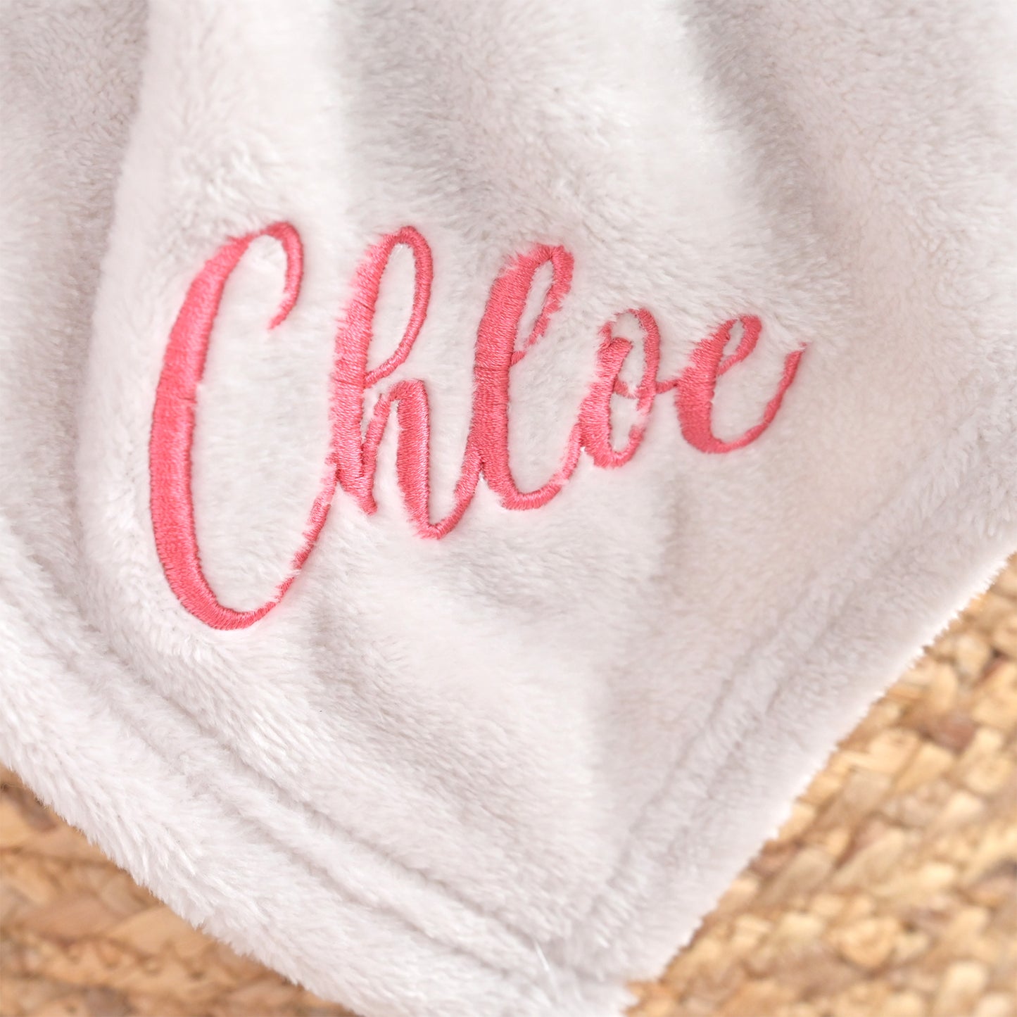 A close-up of a soft white plush blanket with the name 'Chloe' embroidered in pink script, resting on a woven straw surface.