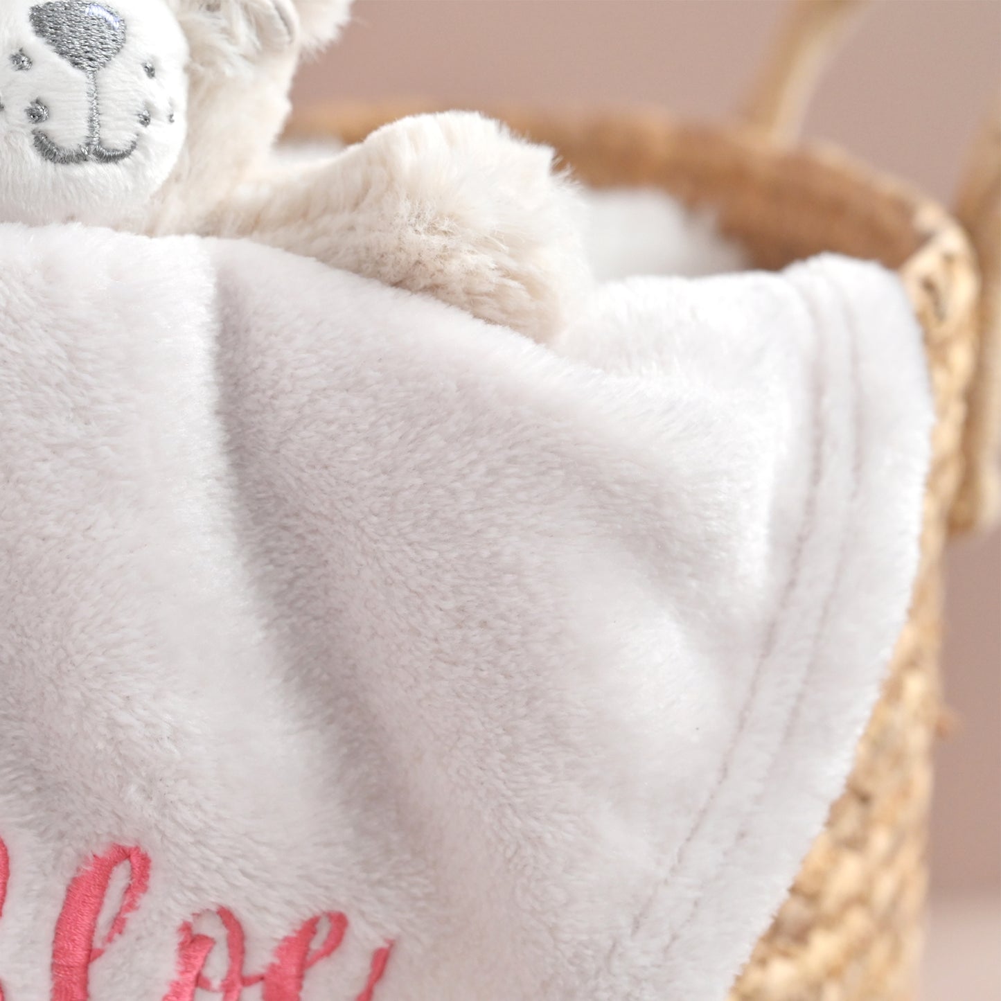 A close-up of a soft white plush blanket draped over a woven basket, with part of a cream stuffed animal visible at the top left and pink embroidered lettering at the bottom.