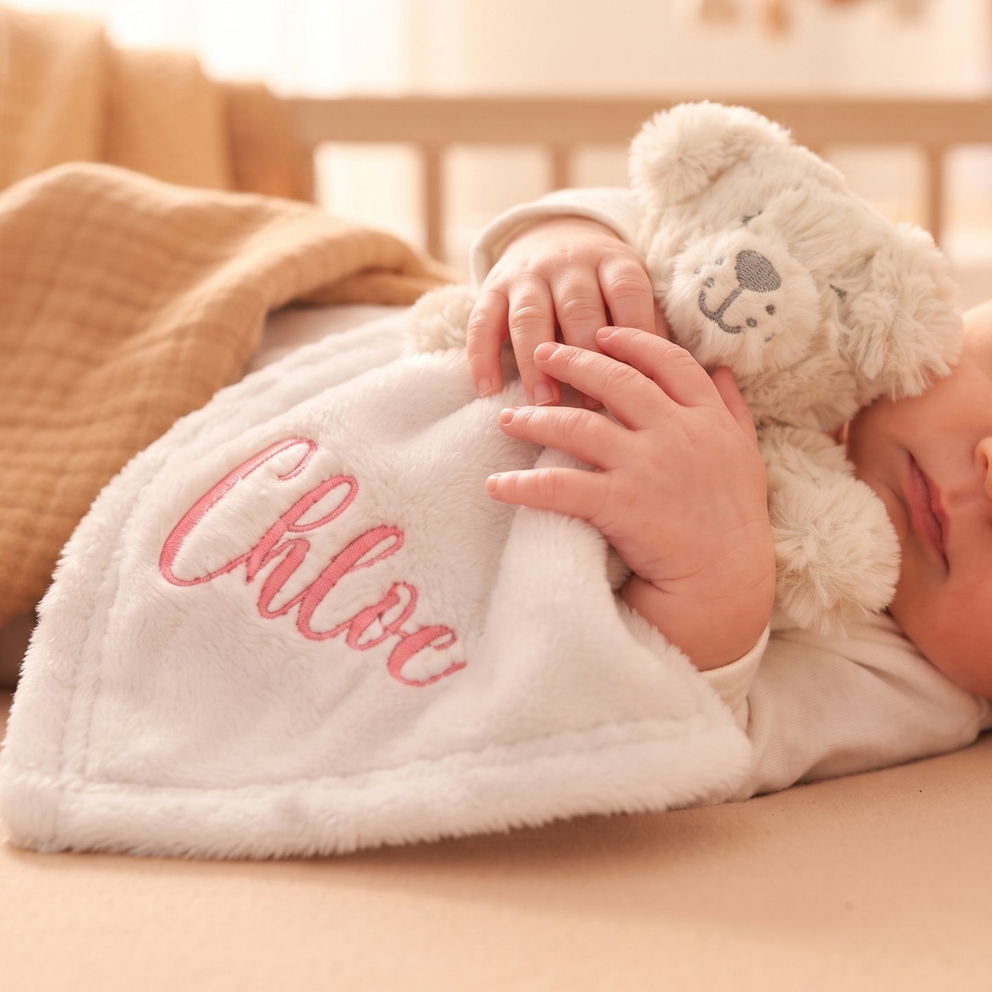 A close-up lifestyle shot of a baby sleeping soundly under a soft white plush blanket embroidered with the name 'Chloe' in pink. The baby is holding a small cream-colored teddy bear, and the scene is set in a warm, softly lit nursery.