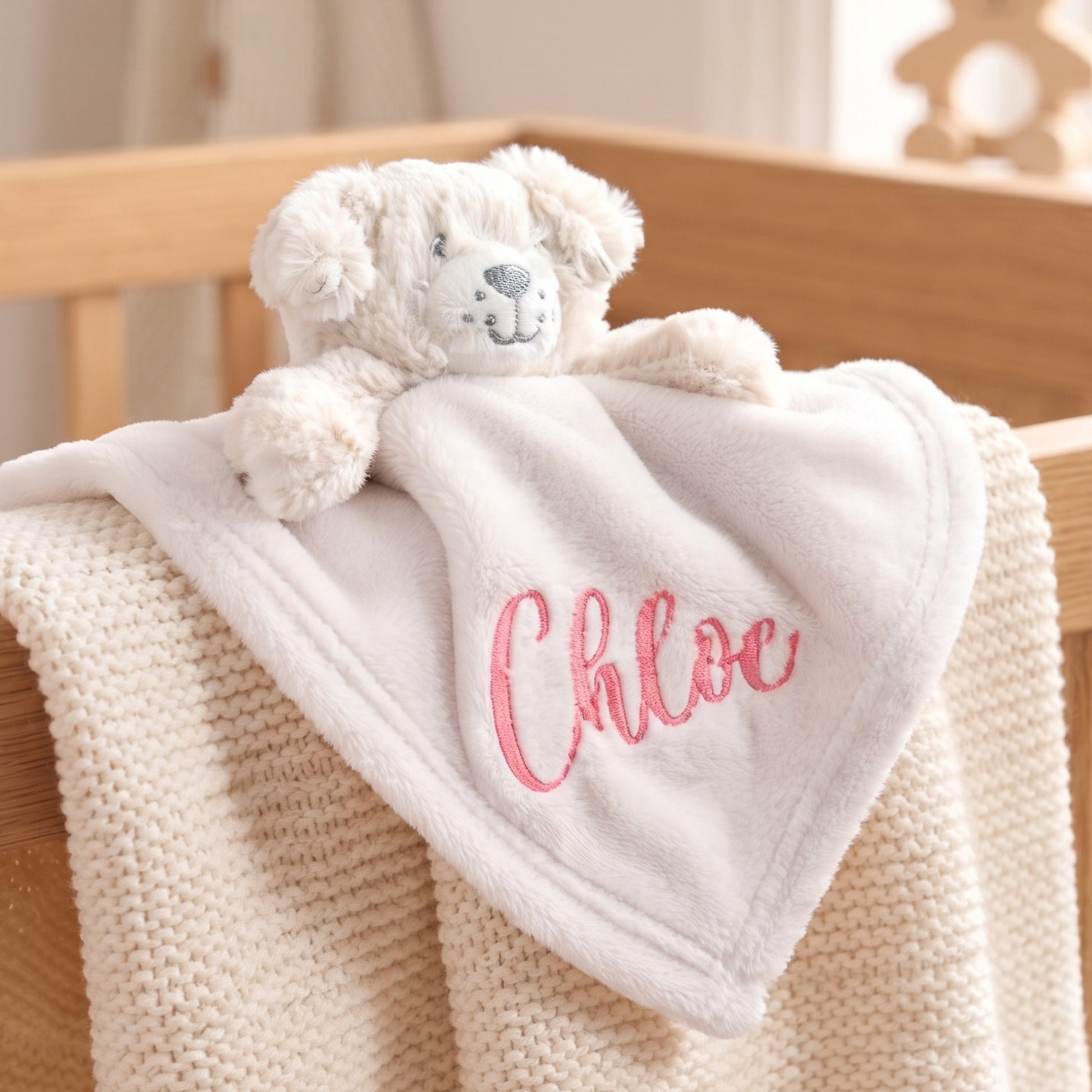 A personalized baby security blanket featuring a plush cream teddy bear attached to a soft white square blanket with the name 'Chloe' embroidered in pink, draped over a beige knit blanket in a wooden crib.