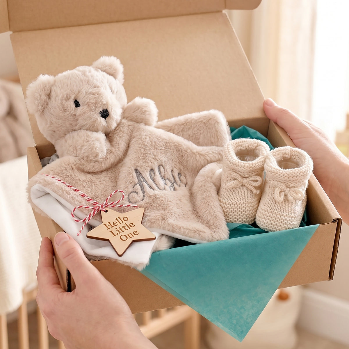 A baby gift set in a cardboard box being held by two hands. The set includes a plush teddy bear, a soft blanket with embroidered text, a wooden star tag reading 'Hello Little One,' and a pair of cream knit booties on a teal tissue paper background.