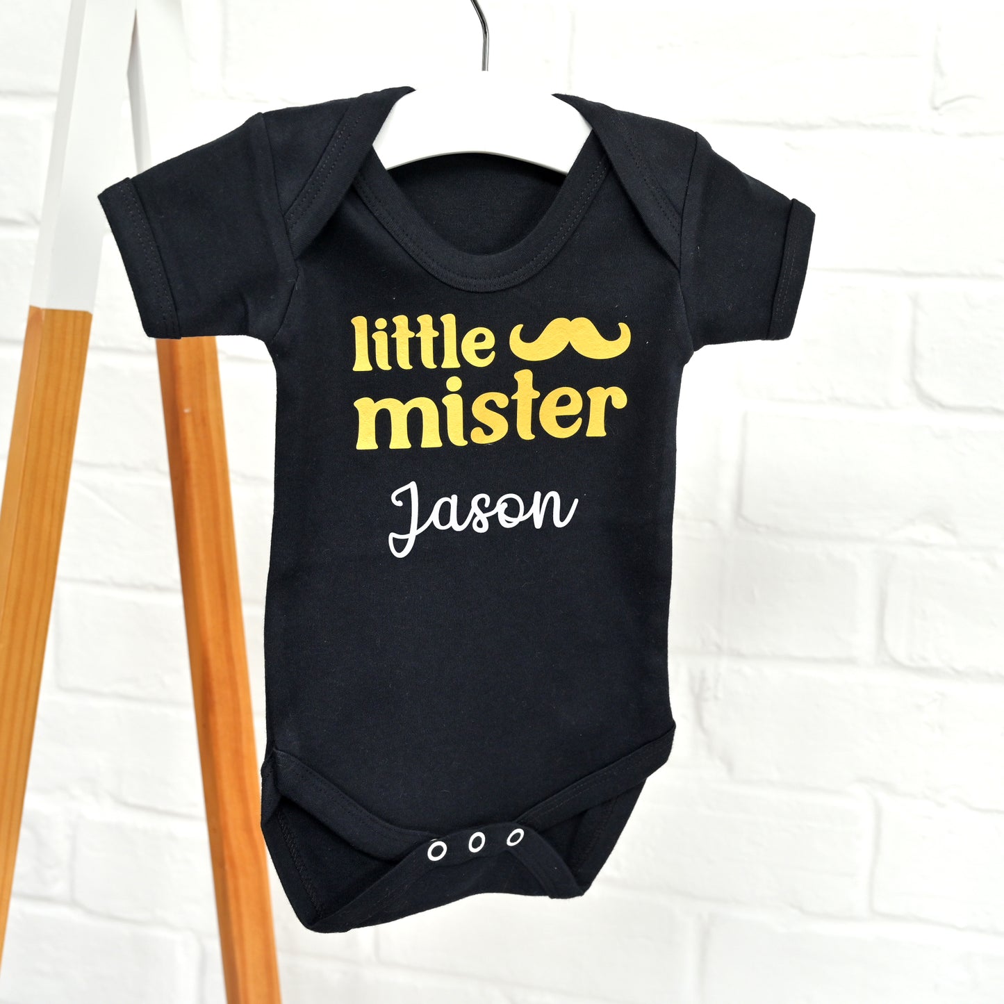 A black short-sleeve baby bodysuit hanging on a white hanger against a white brick wall, with yellow and white printed text reading 'little mister' and 'Jason' plus a yellow mustache graphic.