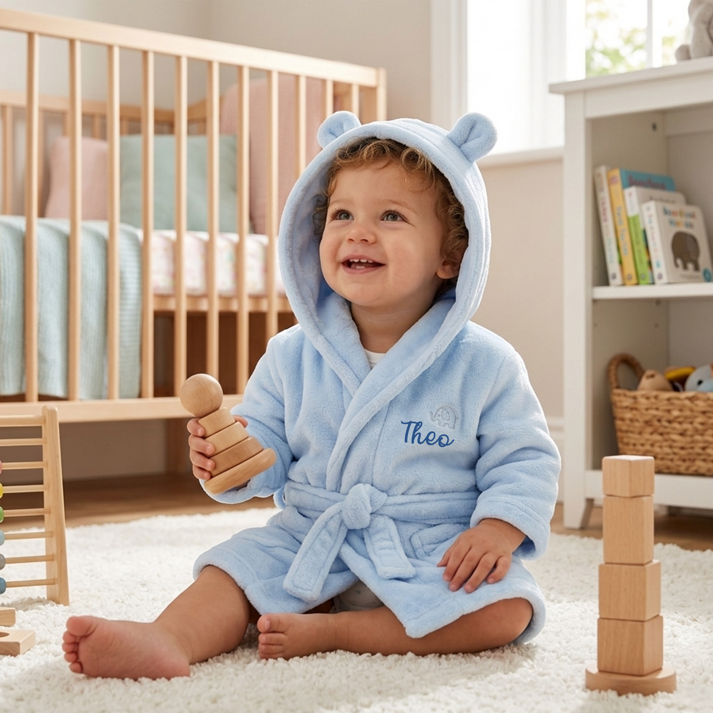 A smiling toddler sits on a white rug in a bright nursery, wearing a light blue plush hooded robe with bear ears and the name 'Theo' embroidered on the chest. The child holds a wooden stacking toy, with a crib, bookshelf, and toys visible in the room.