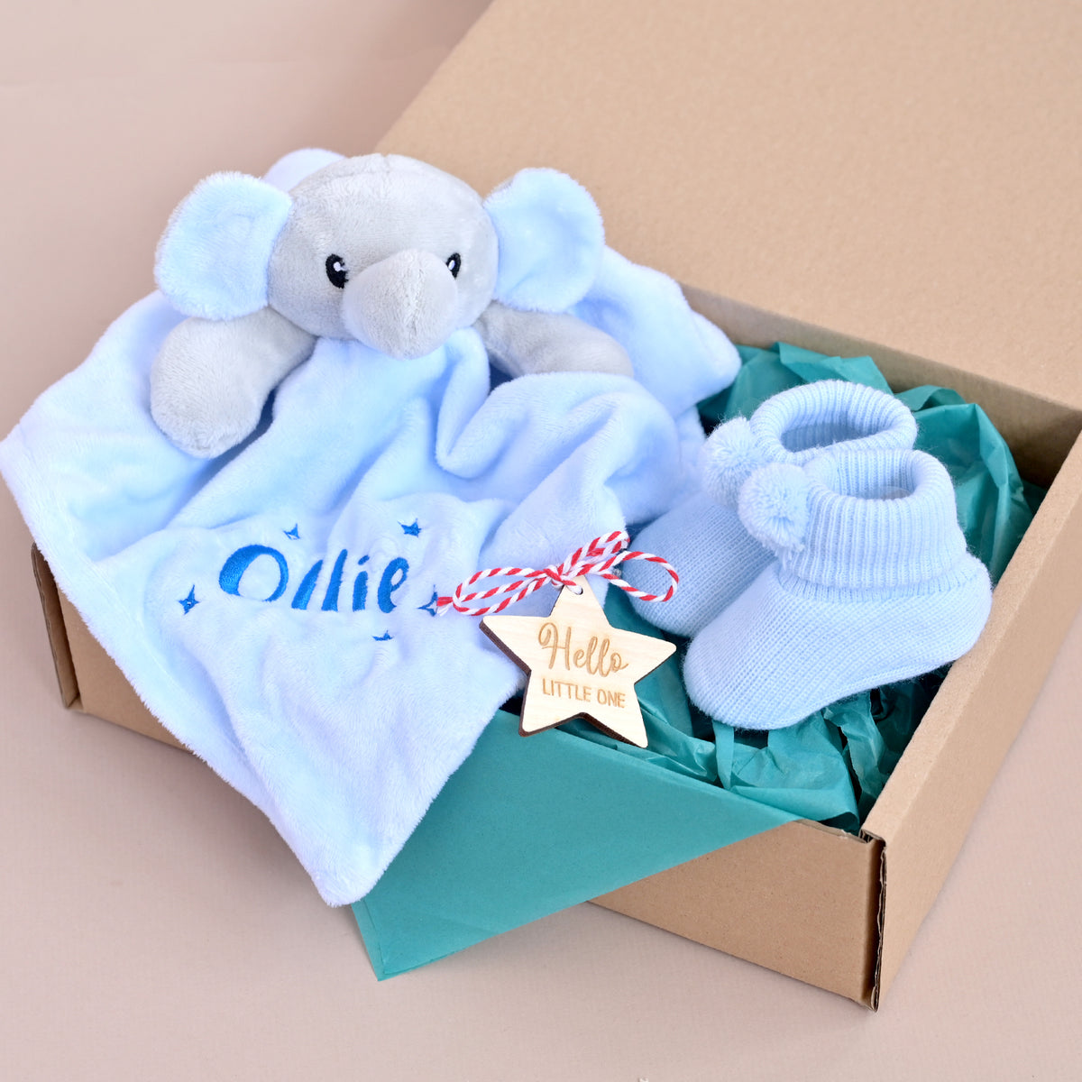 A personalized baby gift set in an open cardboard box, featuring a light blue plush elephant security blanket with 'Ollie' embroidered on it, a pair of light blue knit booties, and a wooden star-shaped tag that reads 'Hello LITTLE ONE'.