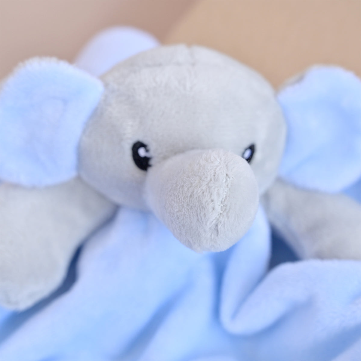 A close-up of a soft plush elephant toy or comforter with a light gray face and trunk, pale blue ears, and a light blue blanket underneath.