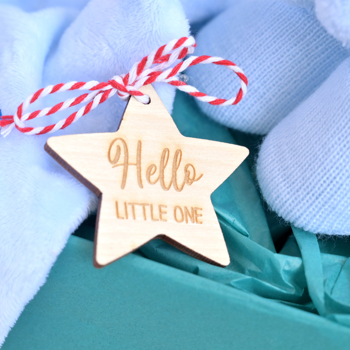 A wooden star-shaped baby keepsake tag engraved with 'Hello LITTLE ONE' in a light natural wood tone, tied with a red and white striped string, nestled in soft blue socks and teal gift tissue paper.