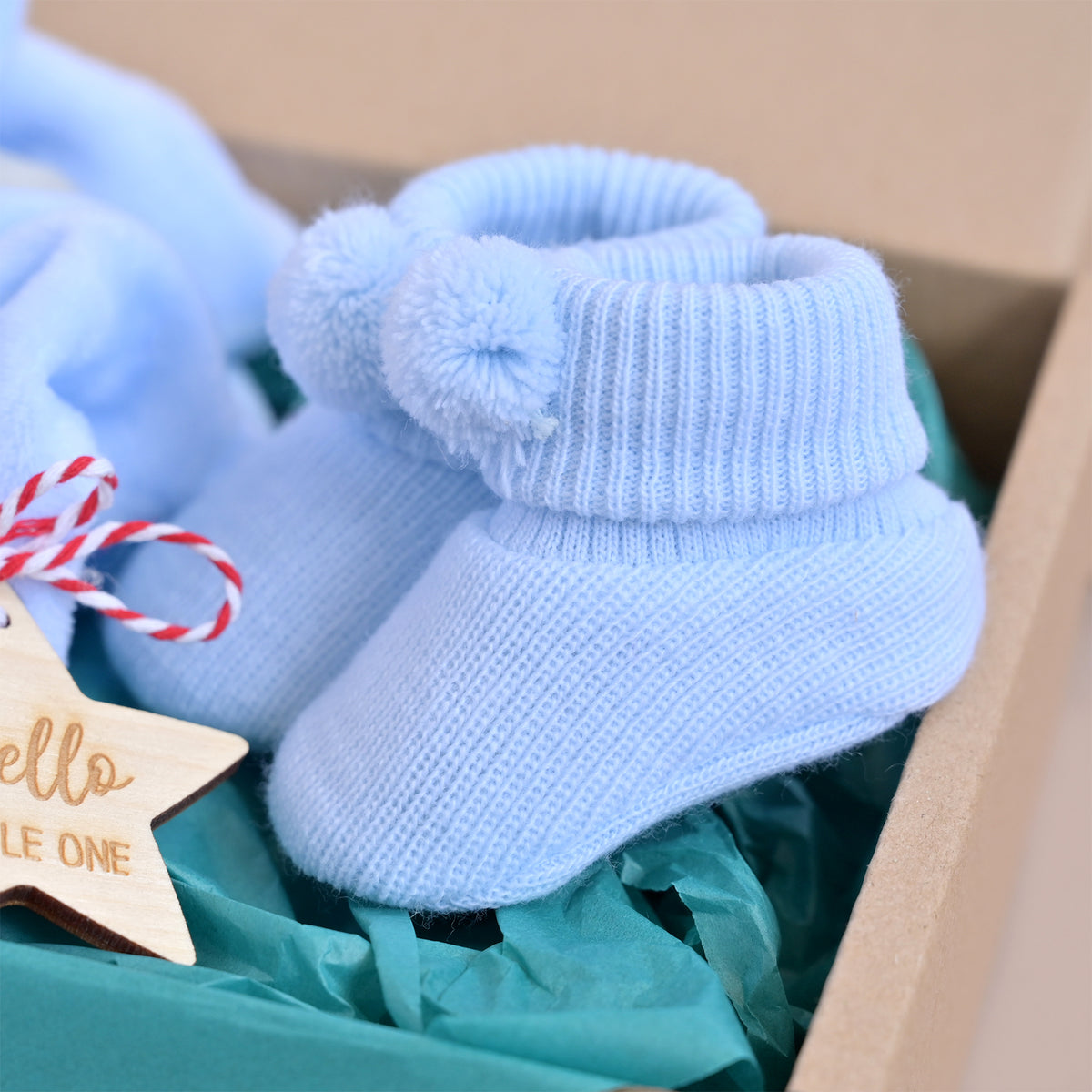 Light blue knit baby booties with fluffy pom-poms on the cuffs, presented in a gift box with teal tissue paper and a wooden star tag.