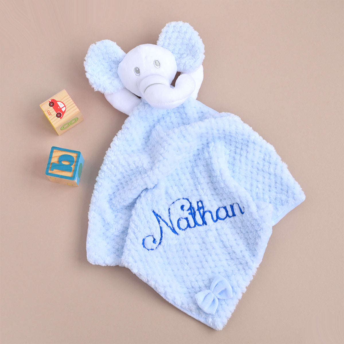 A personalized baby lovey security blanket featuring a plush white elephant head and a textured light blue waffle-weave body with the name 'Nathan' embroidered in blue script. Two small wooden toy blocks are placed beside it on a solid beige background.