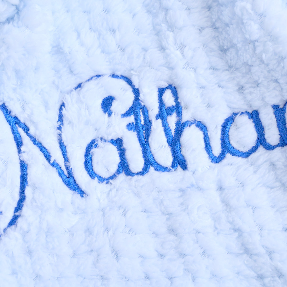 Close-up of a white sherpa fleece blanket with blue embroidered script text reading 'Nathana'.