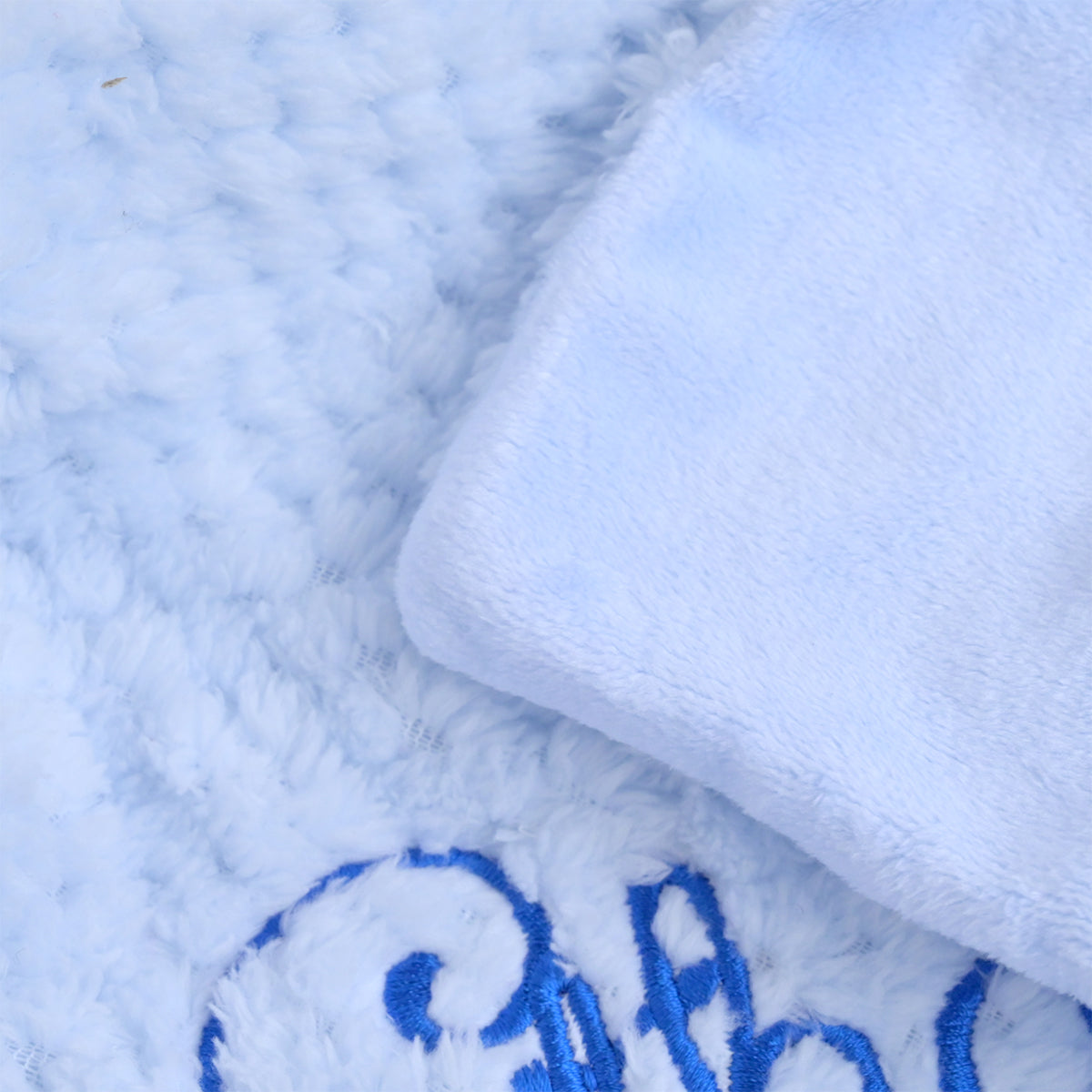 Close-up of a plush light blue blanket or towel with a folded corner showing a soft white underside and blue embroidered lettering.