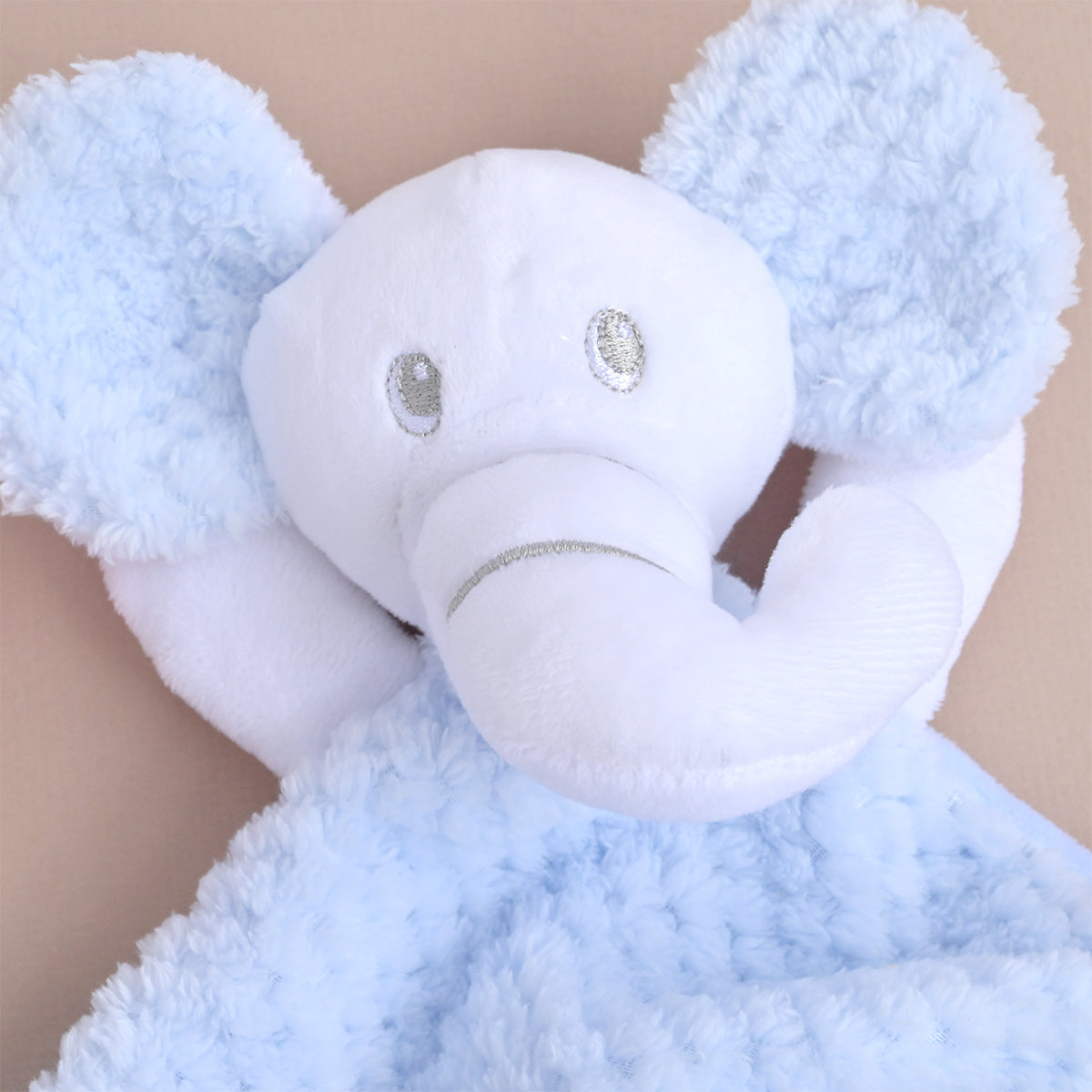 A close-up of a plush elephant security blanket with a soft white head and trunk and fluffy light blue ears and body, shown against a plain beige background.