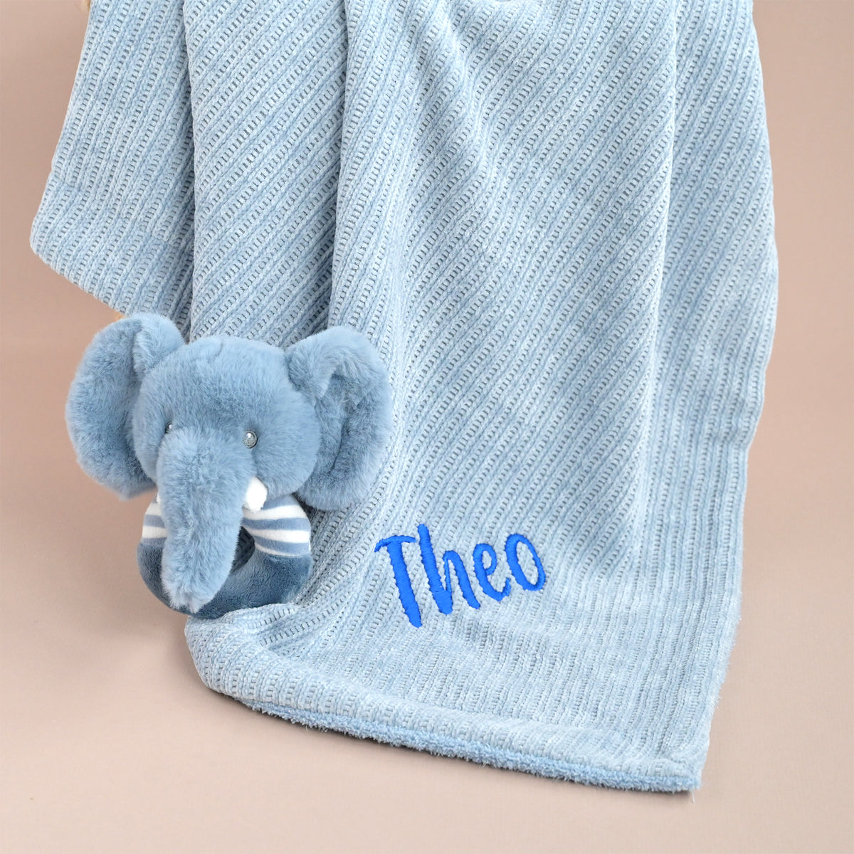 A light blue textured baby blanket draped over a soft beige surface, featuring an embroidered blue name 'Theo' and a small plush elephant toy tucked under the corner.