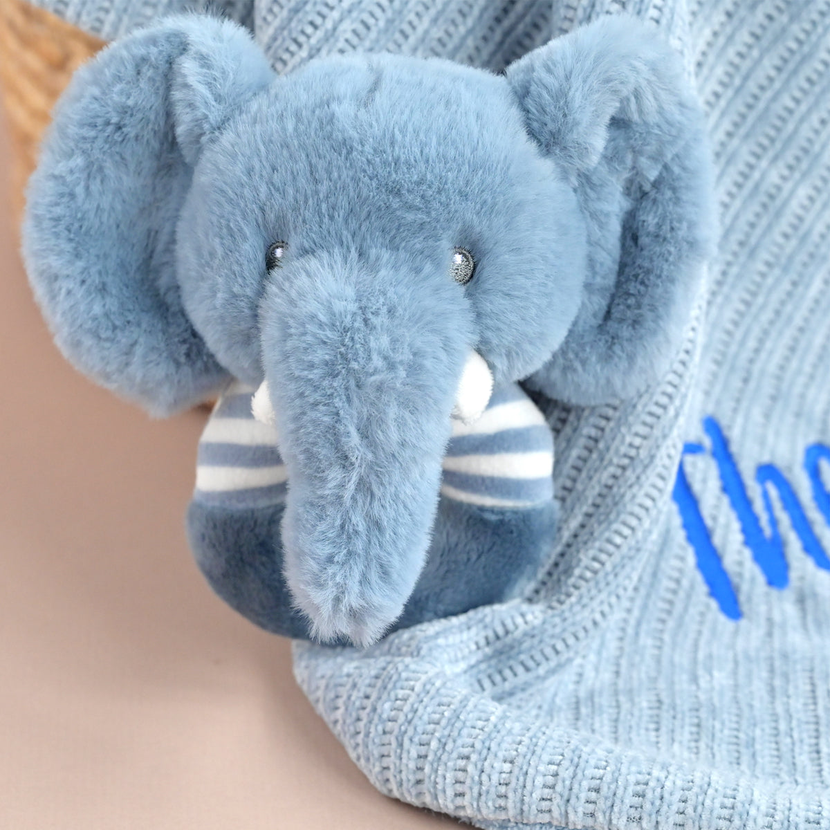 A plush elephant toy in light blue with a white and blue striped scarf, resting against a light blue ribbed knit blanket with blue embroidered lettering.