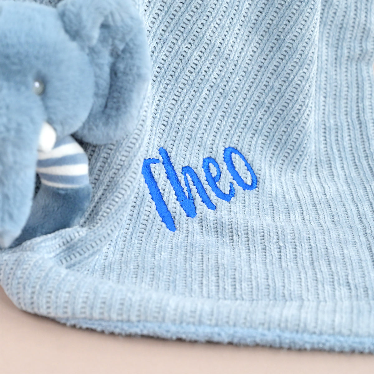 Close-up of a light blue ribbed knit blanket with the name 'Theo' embroidered in bright blue script, next to a plush blue elephant toy.