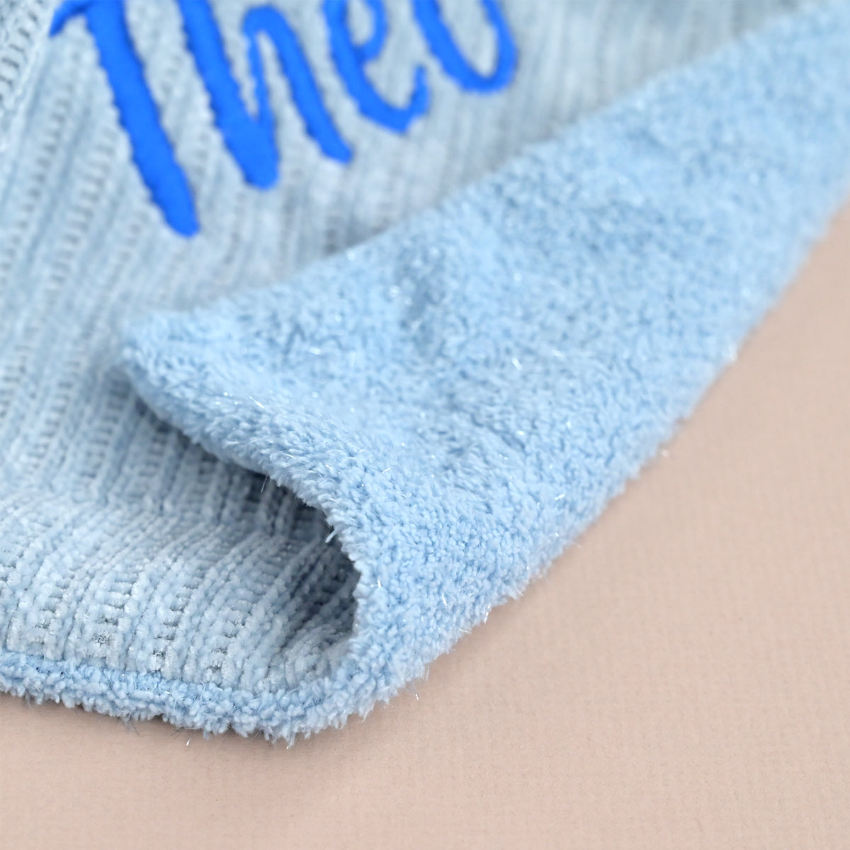 Close-up of a light blue textured fabric with a ribbed knit edge and blue embroidered lettering on a beige background.