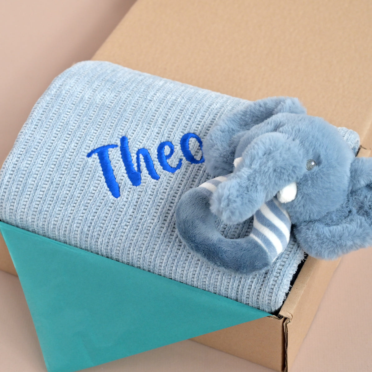 A light blue ribbed knit baby blanket folded inside a brown cardboard box, featuring the name 'Theo' embroidered in blue on the front and a plush blue elephant toy with white stripes resting on top.