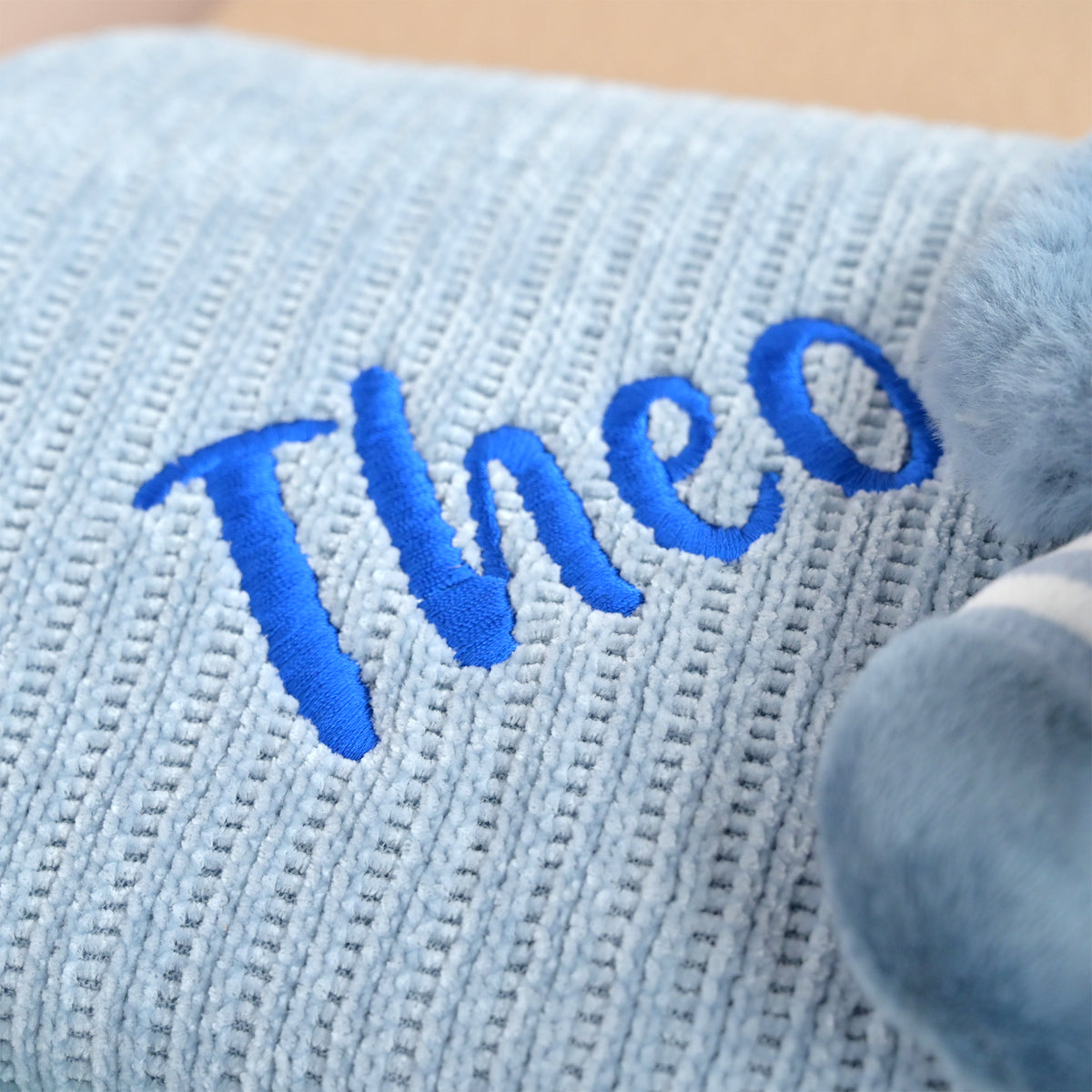 Close-up of a light blue ribbed knit blanket with the name 'Theo' embroidered in bright blue script, next to a soft gray plush toy.