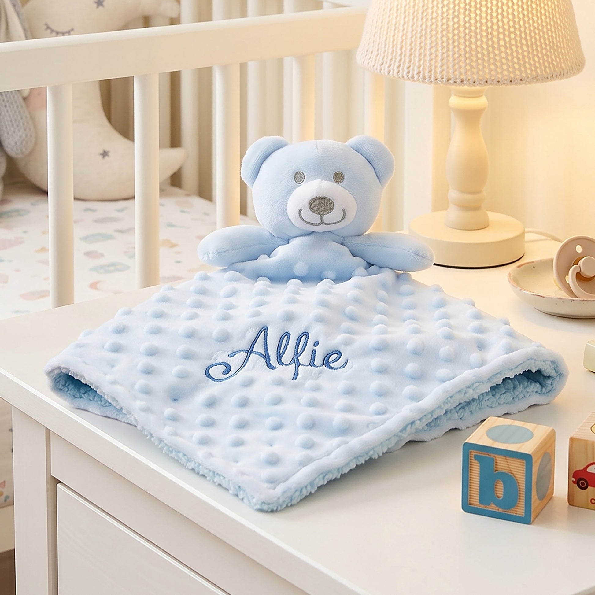 A light blue minky dot comforter with an attached plush teddy bear and the name 'Allie' embroidered on it, styled on a white dresser next to a lamp and baby blocks.