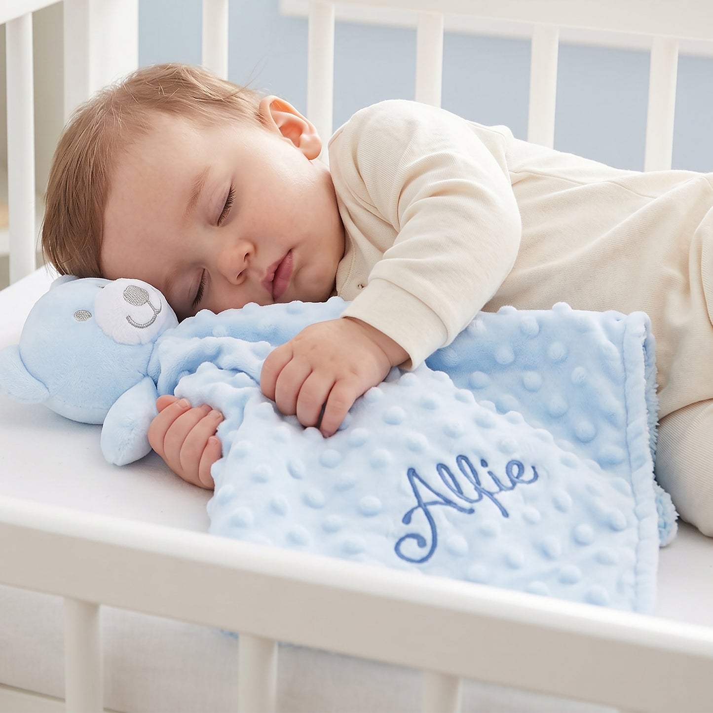 A sleeping baby in a cream onesie rests on a light blue minky dot blanket with a plush bear toy and the name 'Allie' embroidered in blue, placed in a white crib.