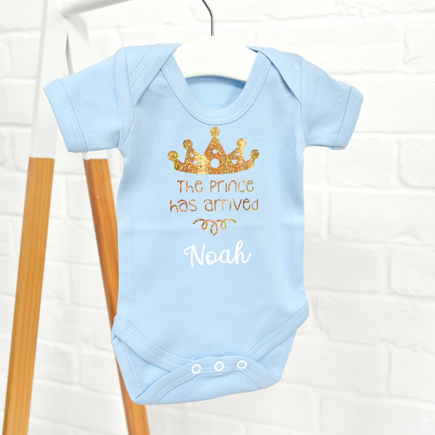 A light blue baby bodysuit hanging on a white hanger against a white brick wall. The bodysuit features a gold glitter crown graphic and the text 'The Prince Has Arrived' and 'Noah' in white and gold lettering.