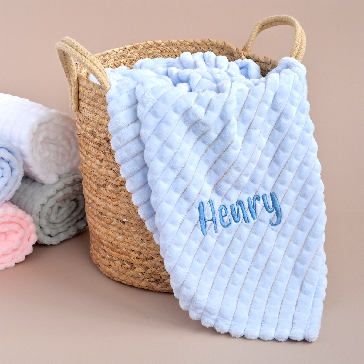 A personalised square blue jacquard blanket with the name 'Henry' embroidered on it, displayed in a wicker basket alongside other folded blankets in pink, grey, and white.