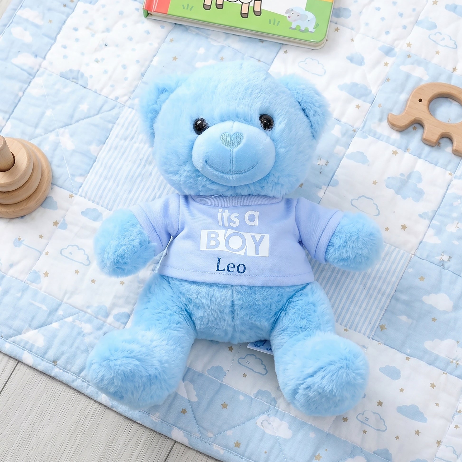 Blue teddy bear soft toy wearing a blue t-shirt printed with "It's a Boy" in bold lettering, personalised with the name Leo, sitting on a cloud-print baby blanket.