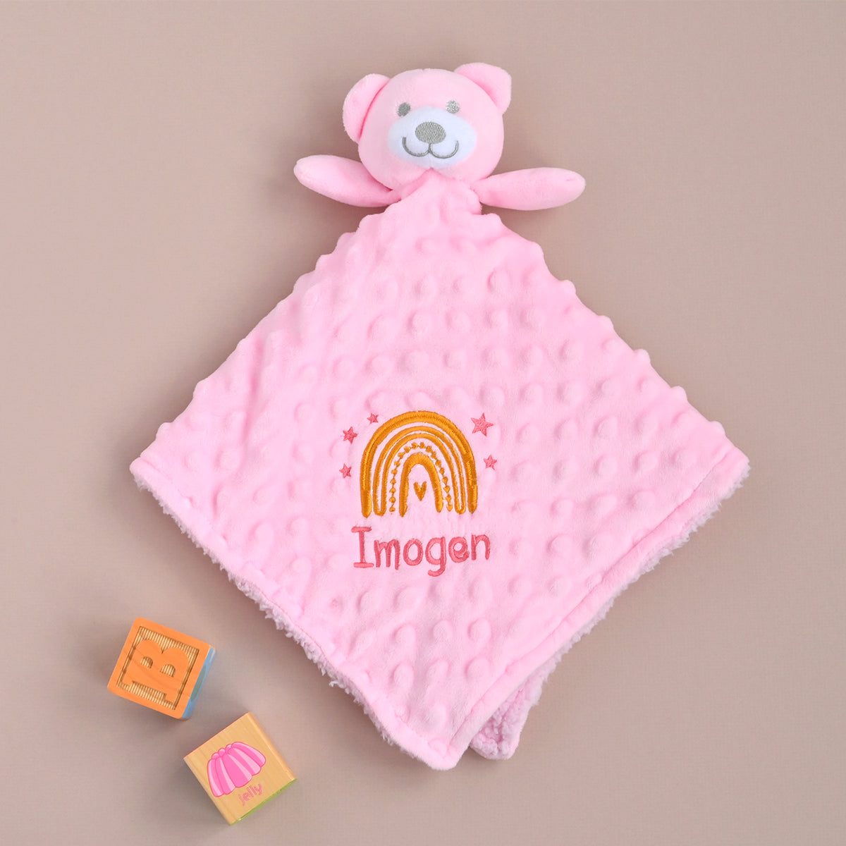A pink minky dot security blanket with a plush bear attachment, featuring a custom embroidered rainbow and the name 'Imogan' on a neutral beige background, with two small toy blocks nearby.