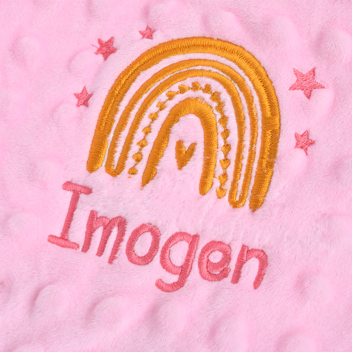 A close-up of a pink textured fabric featuring an embroidered golden-yellow rainbow with pink stars and the name 'Imogen' stitched below in pink.