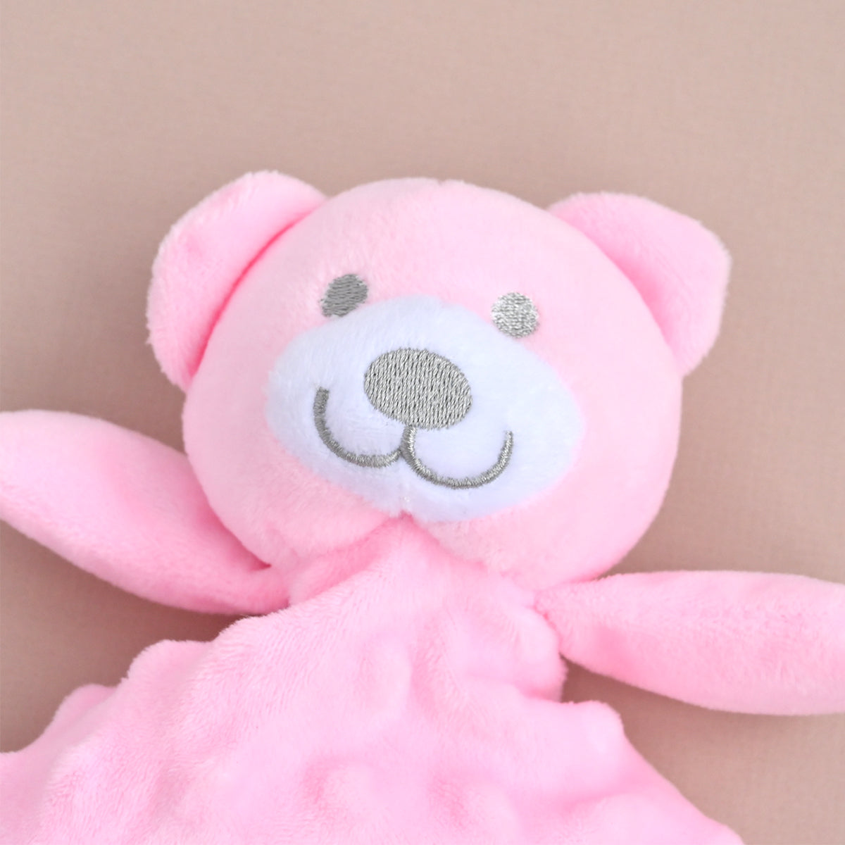 A close-up of a pink plush bear comforter or lovey with embroidered facial features and a textured pink blanket attached, set against a soft beige background.