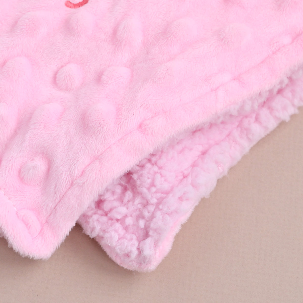 Close-up of a soft pink fleece blanket with a raised dotted texture on the top side and a sherpa-like plush lining on the folded underside, shown on a light beige background.