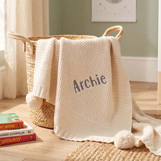 A cream knit baby blanket with the name 'Archie' embroidered in gray, draped over a woven basket in a styled nursery setting.